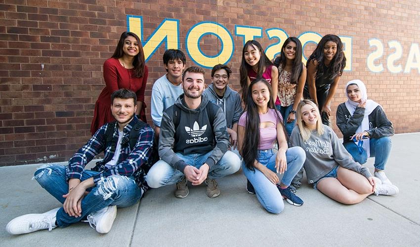 students seated on ground in a tight group in front of a UMass Boston sign on brick wall