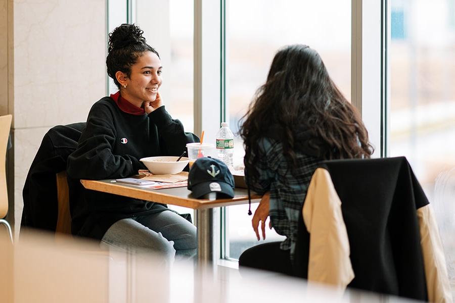 two female students talk casually at seated window in UMass Boston Campus Center Dining Hall
